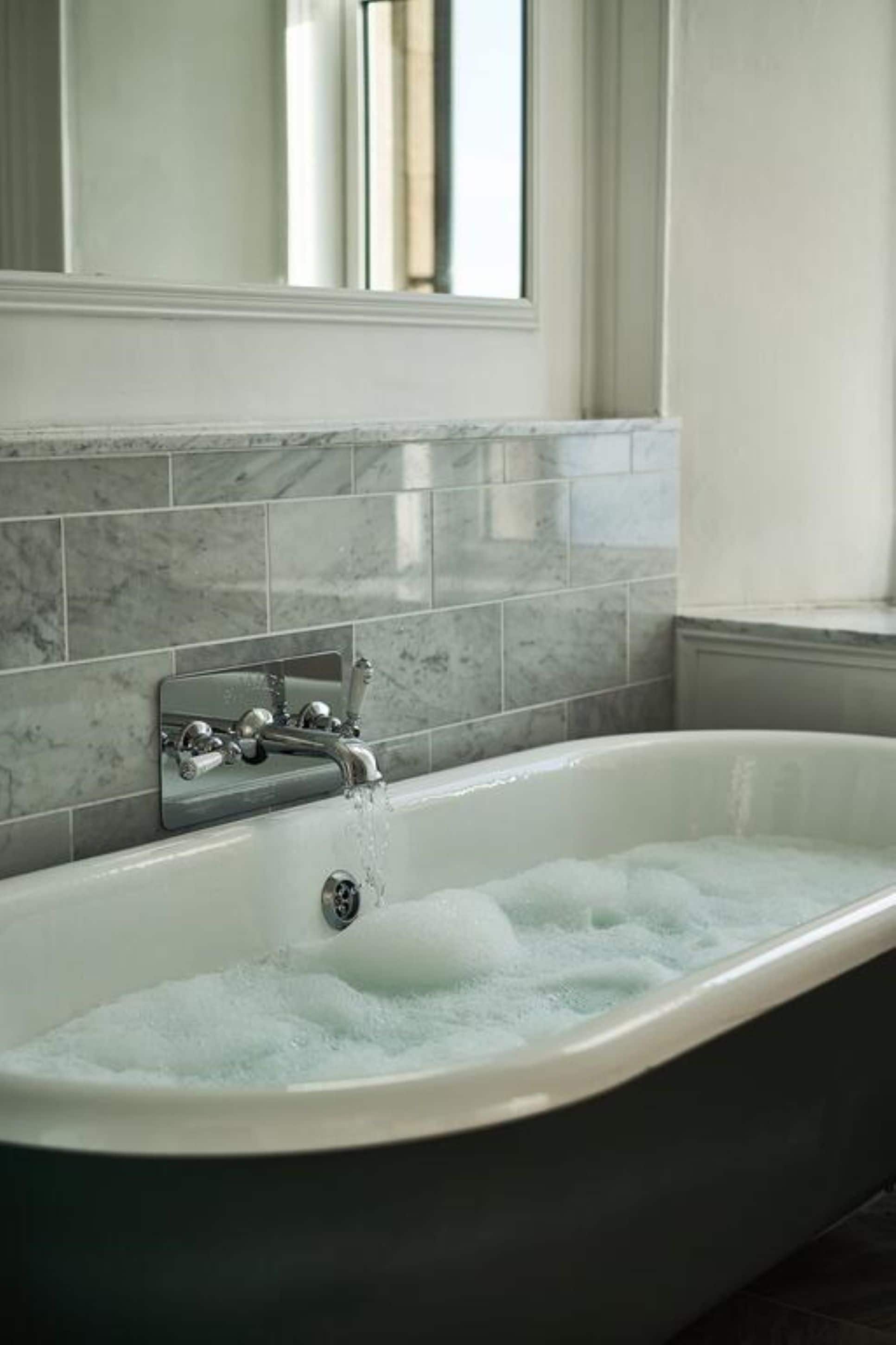 Un baño moderno con una bañera independiente, un espejo encima y azulejos de mármol en la pared.
