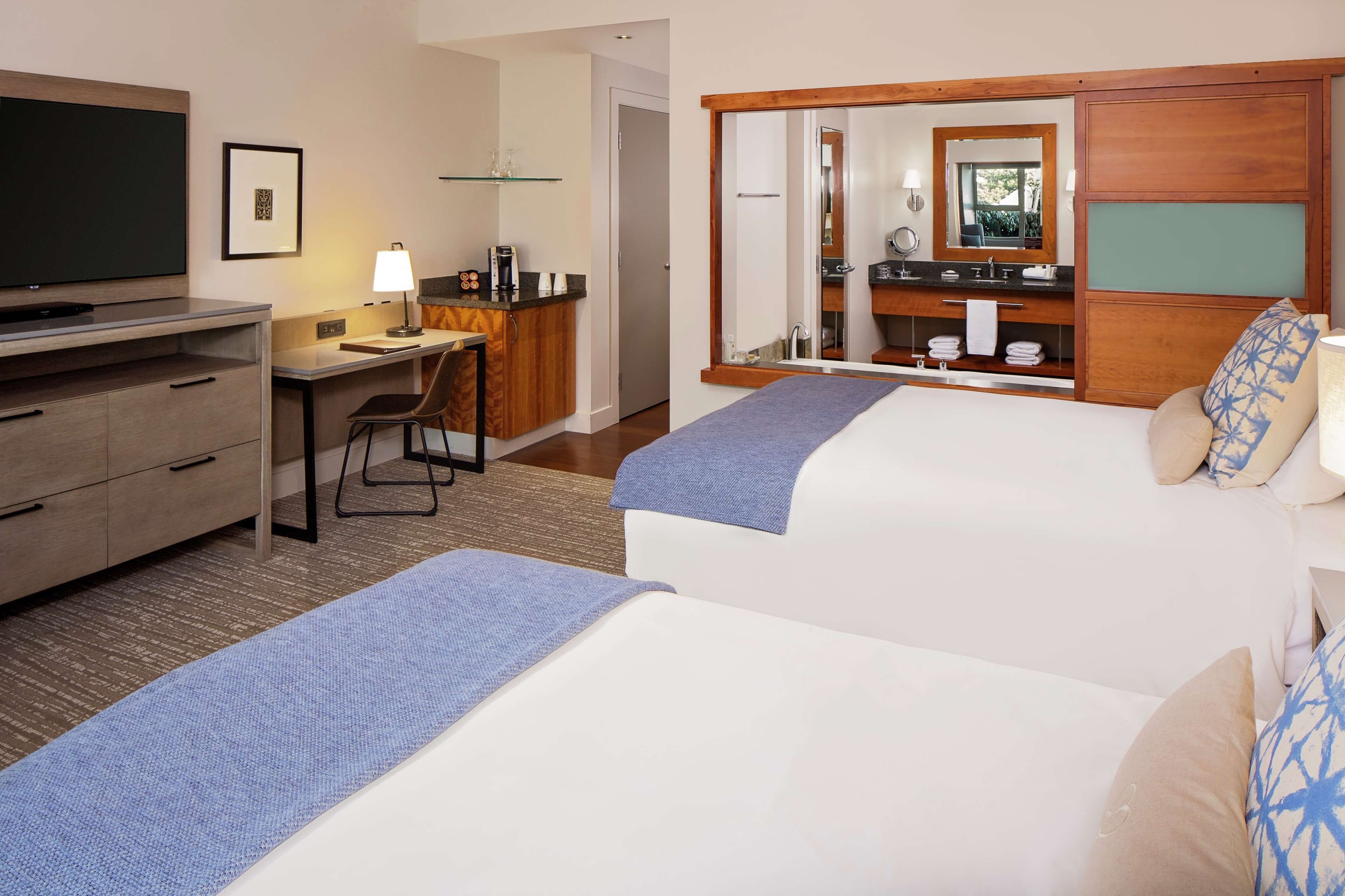 A hotel room with two beds, a desk with a chair, a television, a small kitchenette, and a bathroom with a mirror.