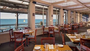 Un restaurant avec des tables dressées pour le service, de grandes fenêtres donnant sur la mer et un menu écrit à la craie.