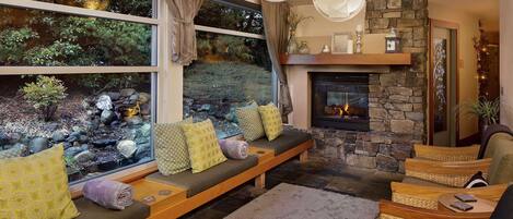 A cozy living room with a stone fireplace, a built-in bench with cushions, and a view of a garden through large windows.