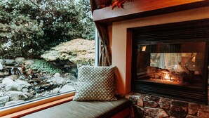 Une cheminée en pierre avec un feu, un banc agrémenté d’un coussin et une vue sur un jardin avec des rochers et des arbres.