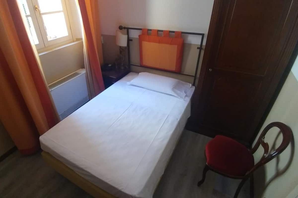 A small, single-bed room with a wooden headboard, a red chair, and a window with curtains.