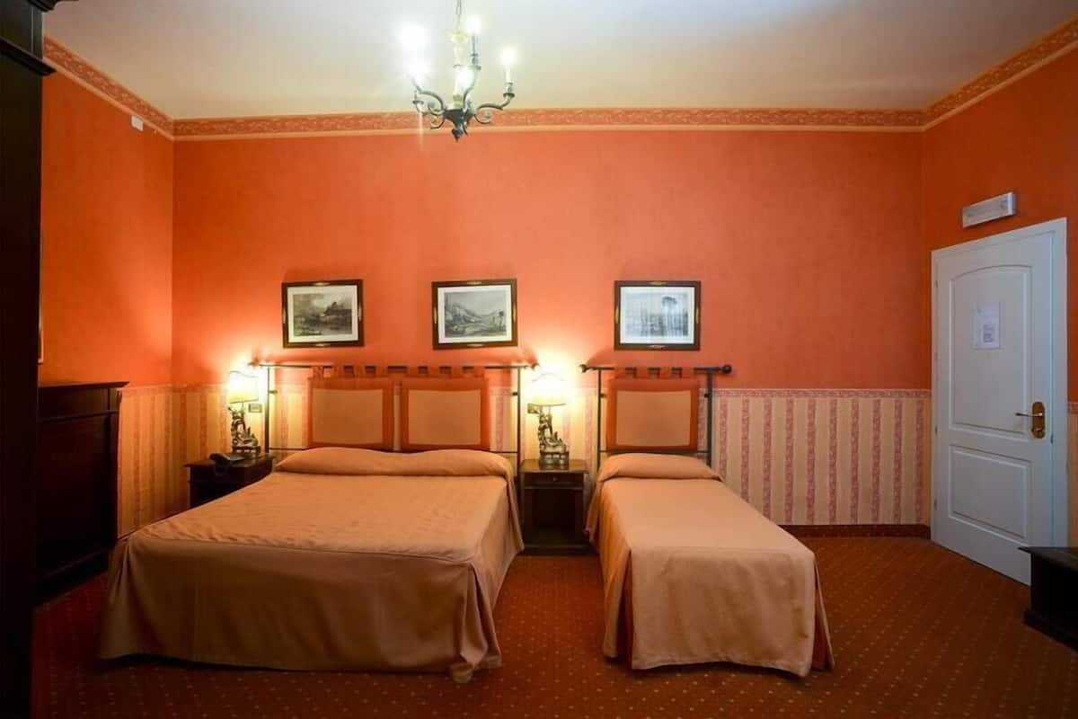 A room with two beds, a chandelier, framed pictures on the wall, and a door.