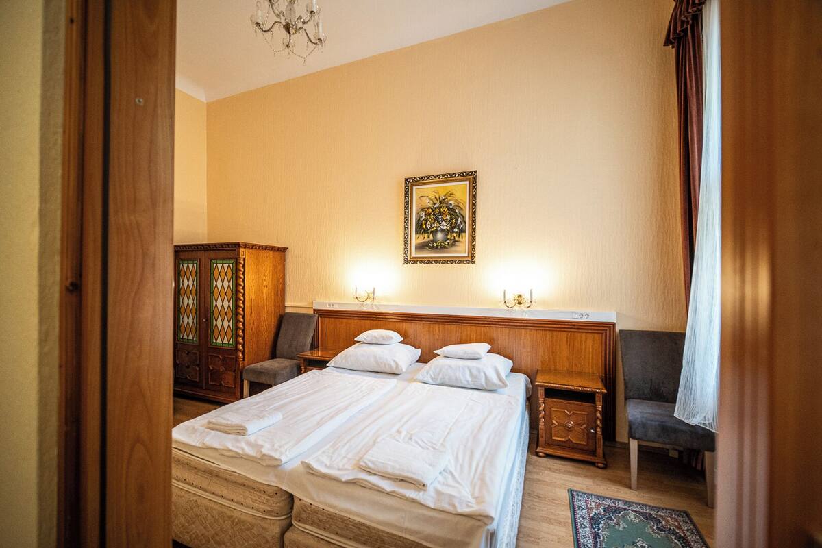 Una habitación de hotel con cama, dos mesitas de noche, un armario de madera, una silla y una araña.