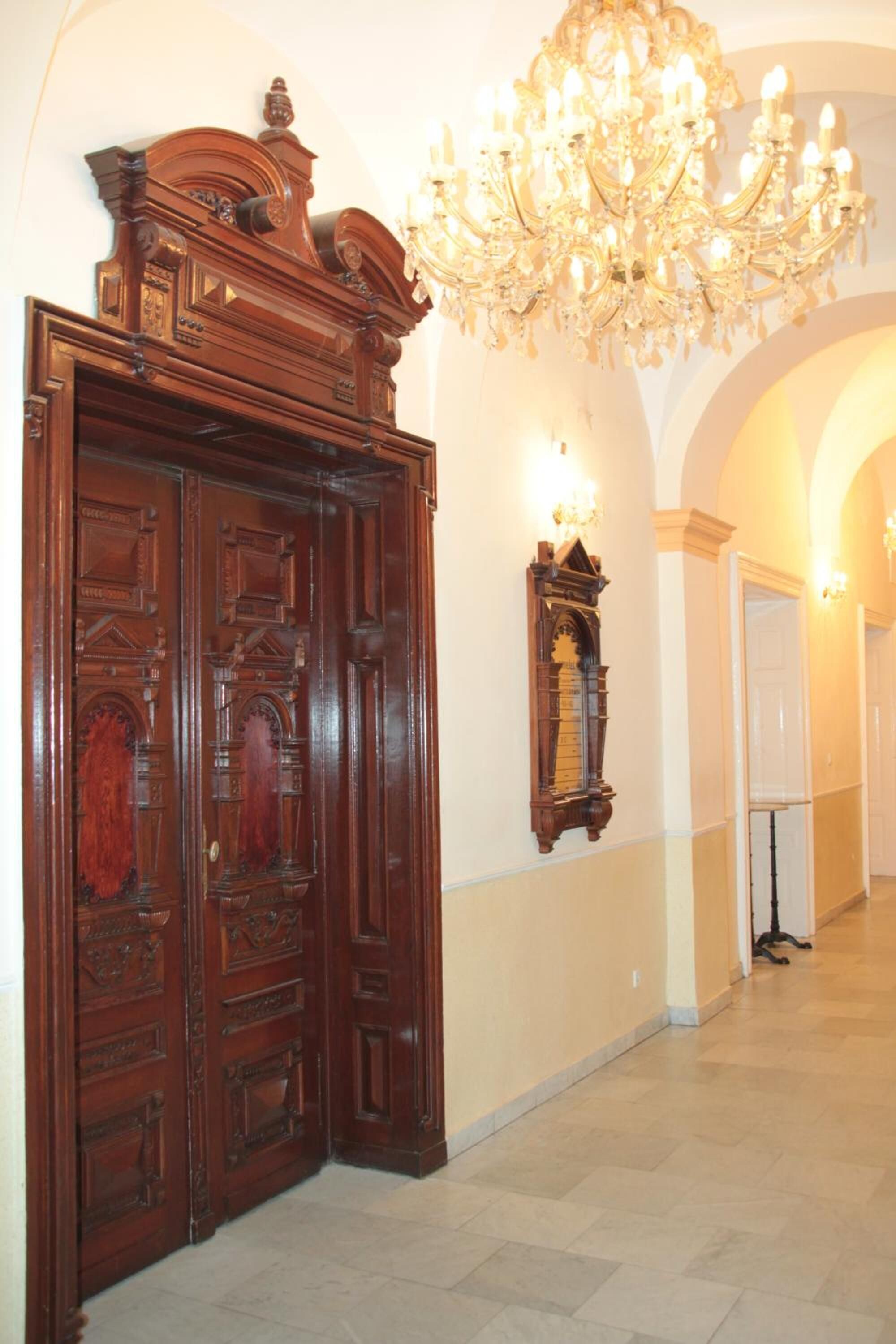 A hallway with a grand wooden door, a wall-mounted clock, and a chandelier.