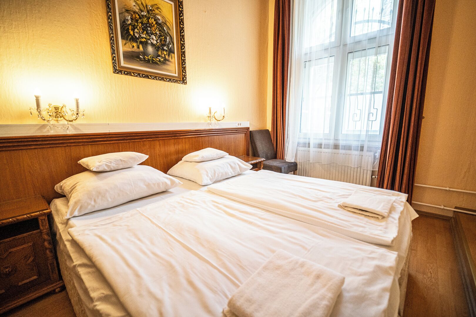A hotel room with a large bed, two bedside tables, a chair, a wall-mounted lamp, and a window with curtains.
