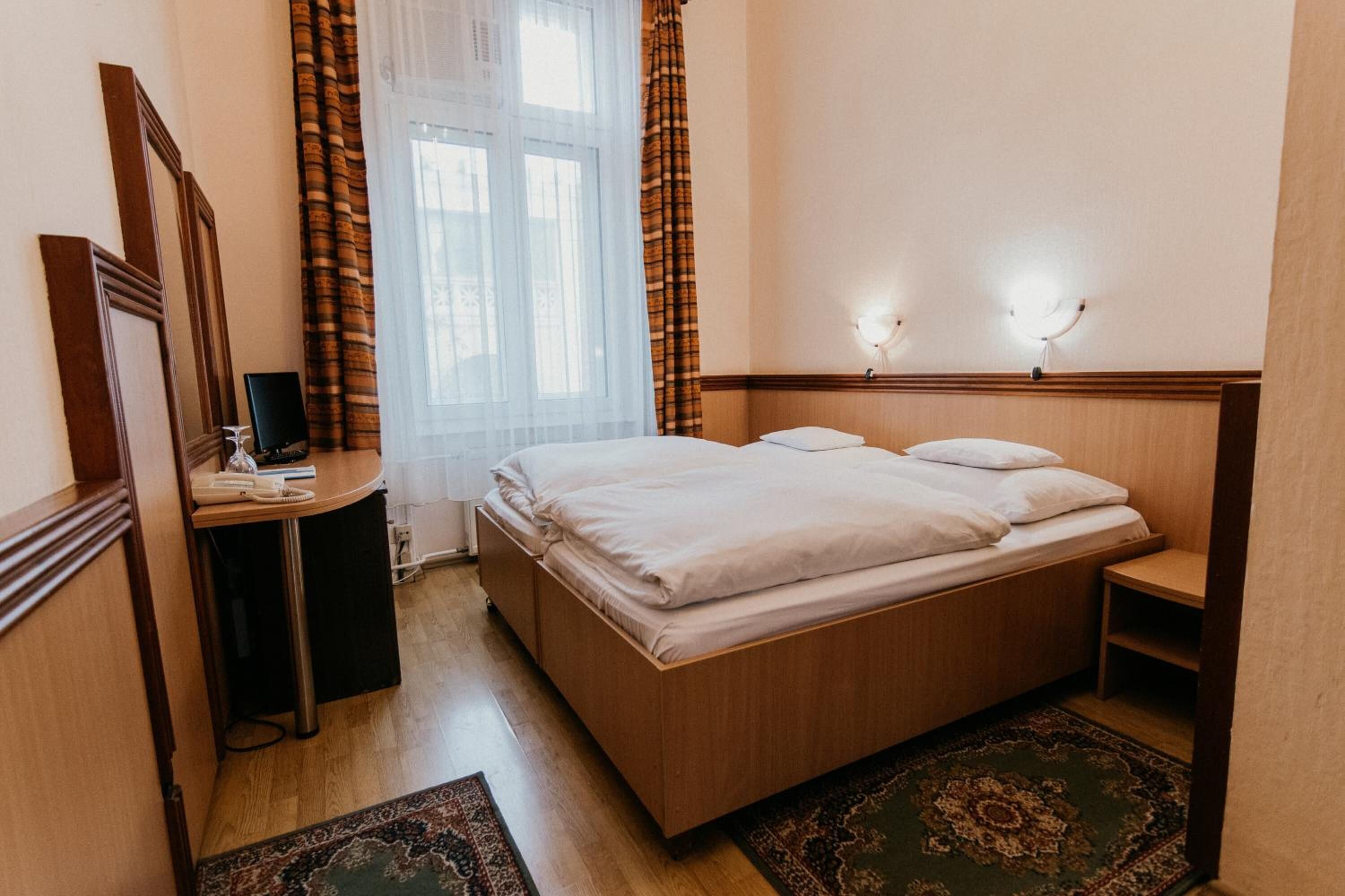 A hotel room with a wooden bed, a desk with a chair, a mirror, and a window with curtains.
