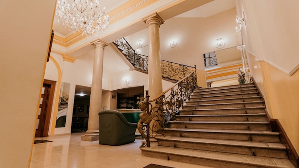 A grand staircase with ornate metal railings, a chandelier, and a green upholstered chair. - Tisza Hotel (Szeged)