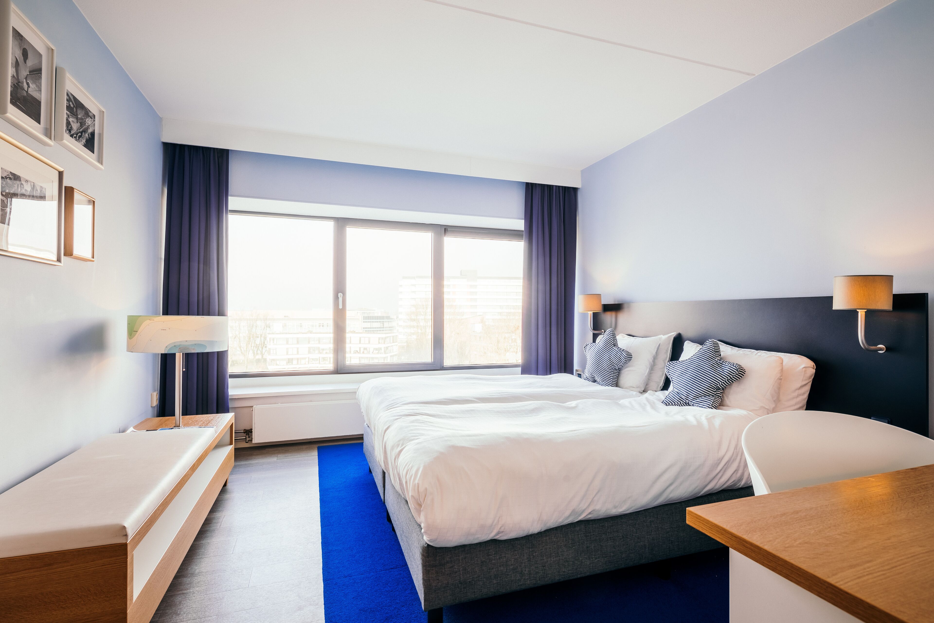 Una habitación de hotel moderna con una cama grande, mesitas de noche, un escritorio y un ventanal con cortinas.