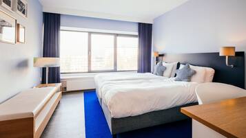 Una habitación de hotel moderna con una cama grande, mesitas de noche, un escritorio y un ventanal con cortinas.