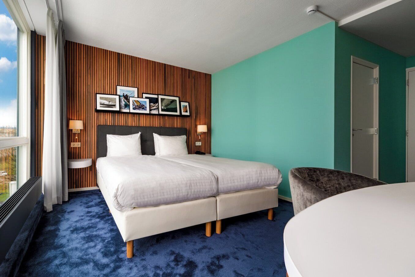 A hotel room with a large bed, a wooden accent wall, a teal-colored wall, and a window with a city view.