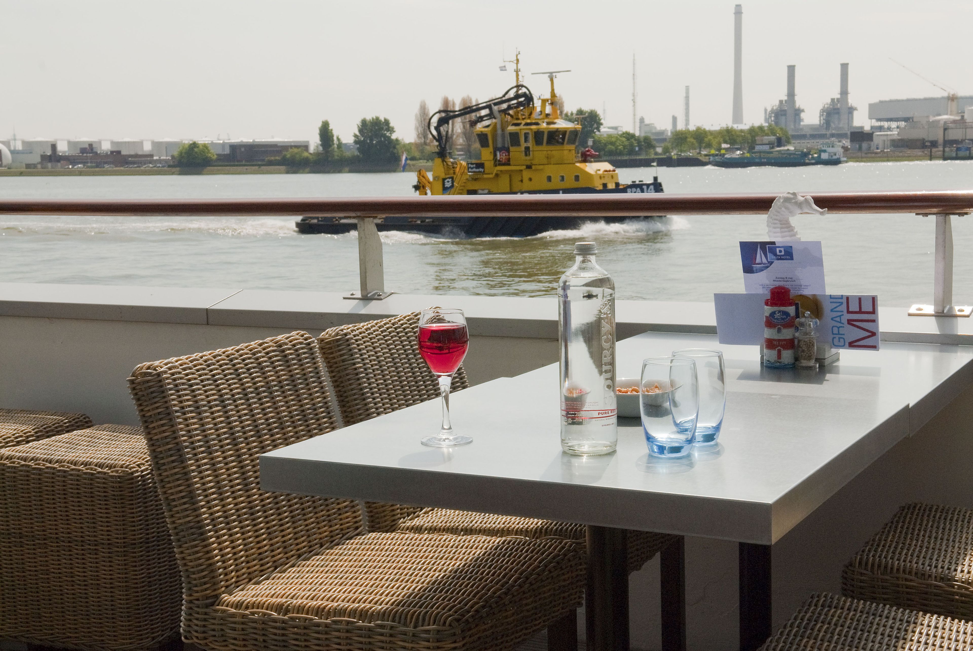 Une table avec une bouteille, des verres et un menu, placée sur un balcon donnant sur une rivière, avec un bateau jaune visible au loin.