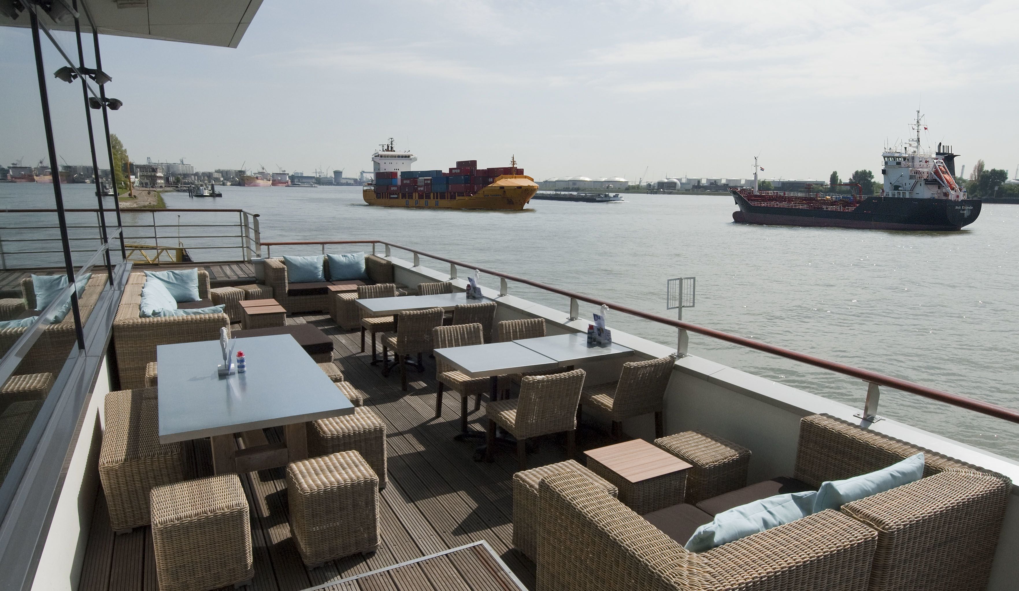 A deck with wicker furniture and tables, overlooking a body of water with large cargo ships.