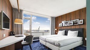 Habitación de hotel con cama, escritorio, silla y un ventanal con vistas a un puente y al agua.