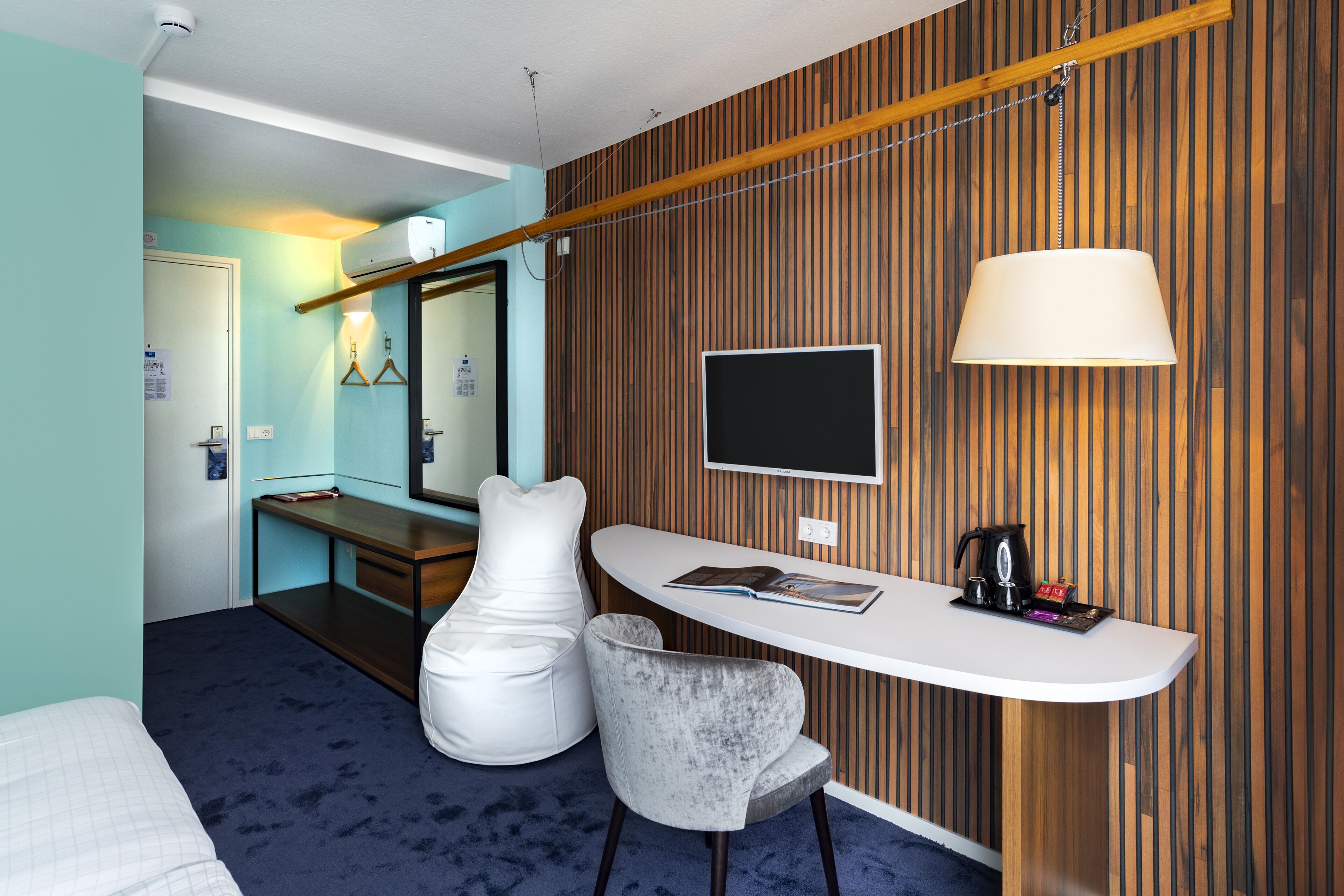 A modern hotel room with a bed, a desk with a chair, a TV, and a wooden accent wall.