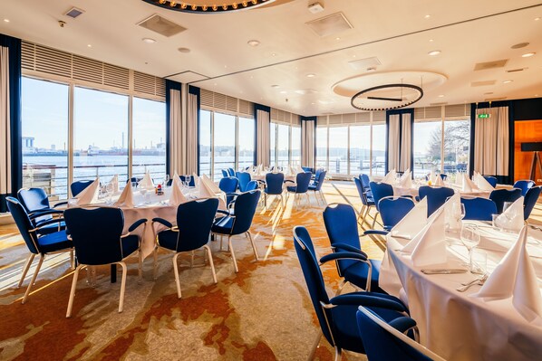 Un salón de conferencias con mesas redondas, sillas azules y grandes ventanales con vistas a un río.