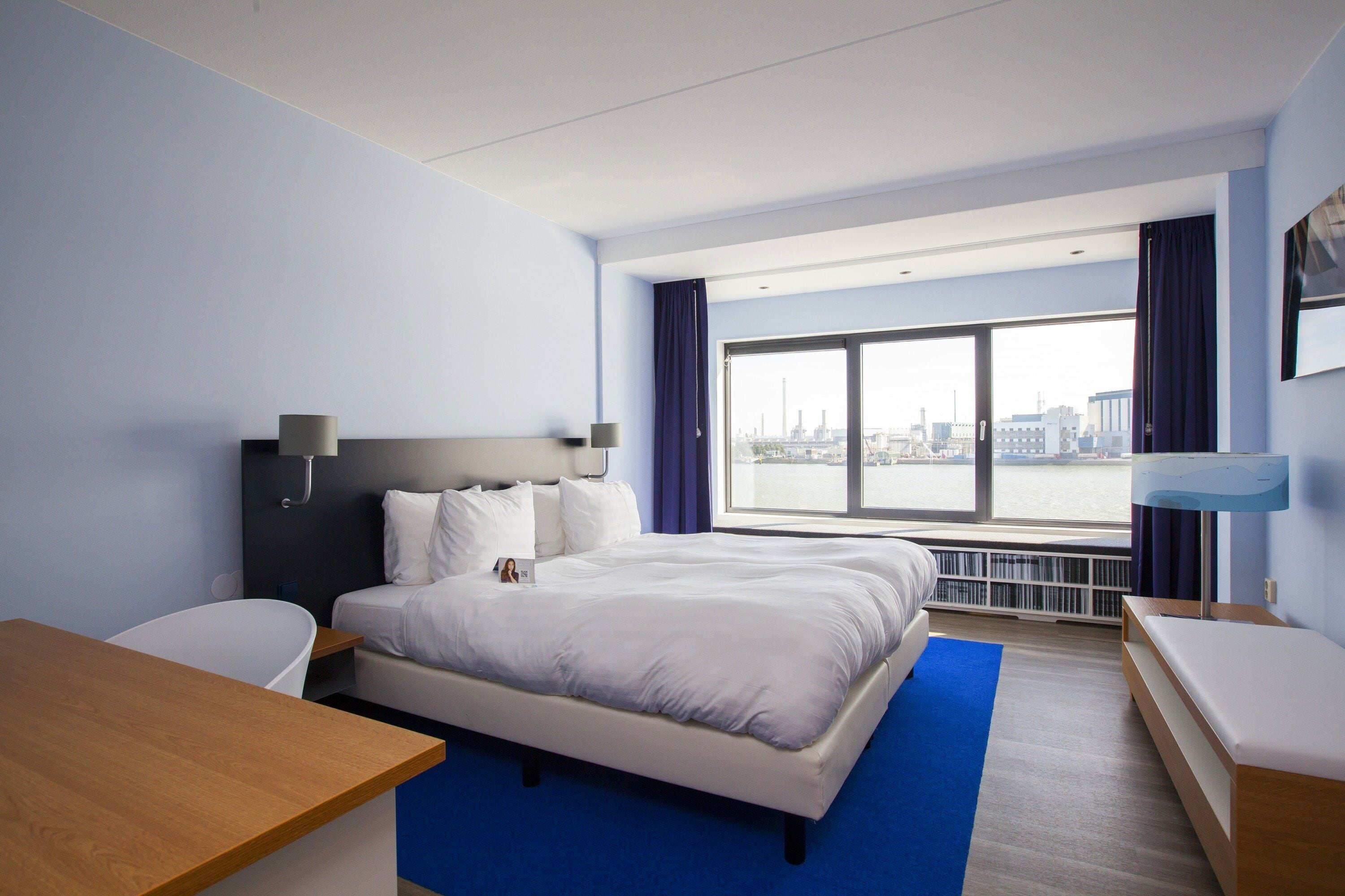 A modern hotel room with a large bed, a desk, and a view of the city through a large window.