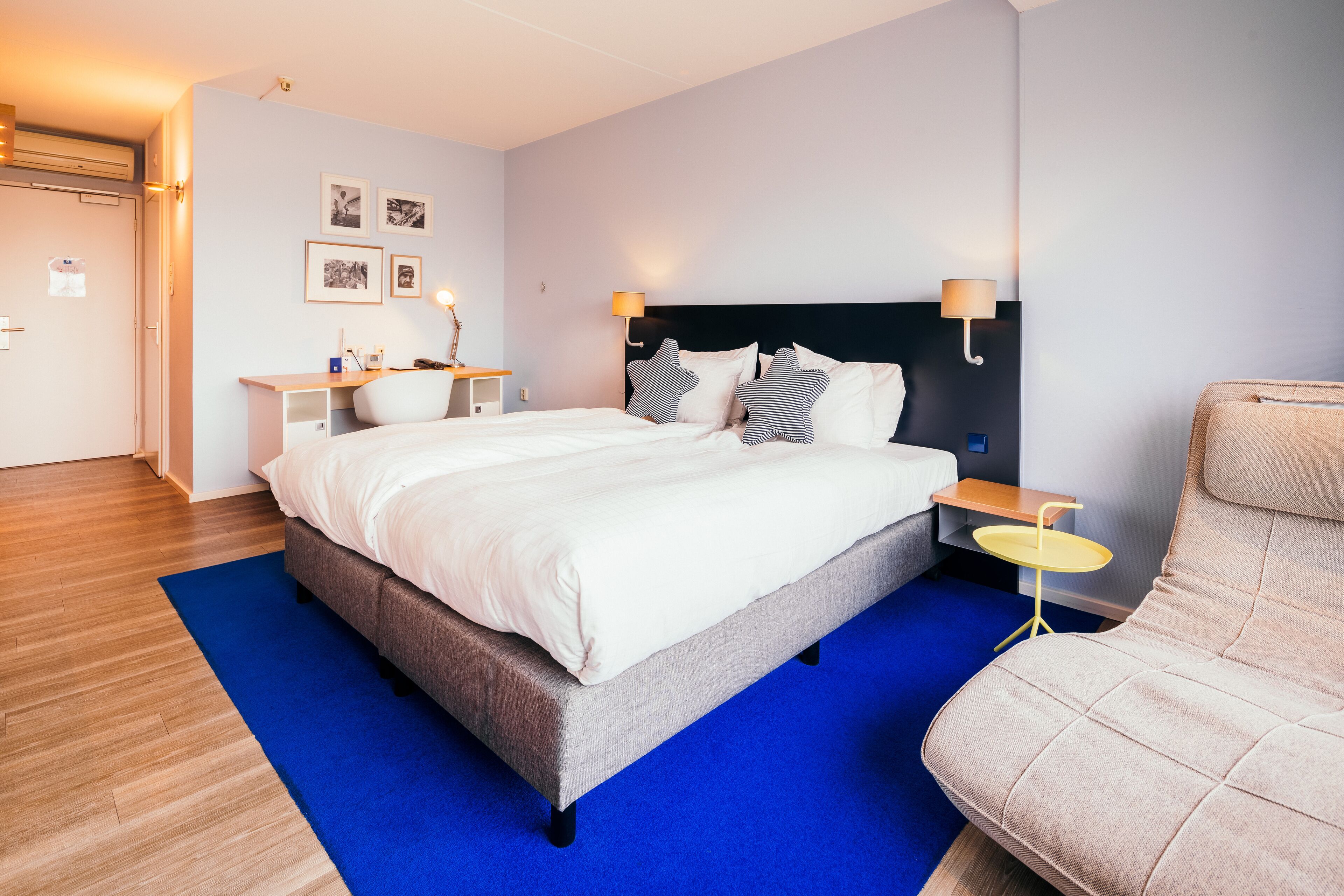 A modern hotel room with a large bed, a bedside table, a chair, and a blue rug.