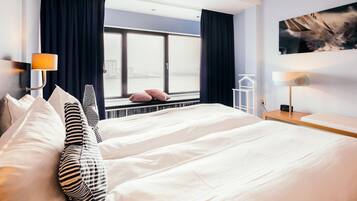 A hotel room with a large bed, a view of the city through a window, and a painting on the wall.