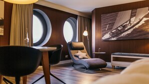 Habitación de hotel moderna con un ventanal grande, una silla de descanso, una mesita y un cuadro de un velero.