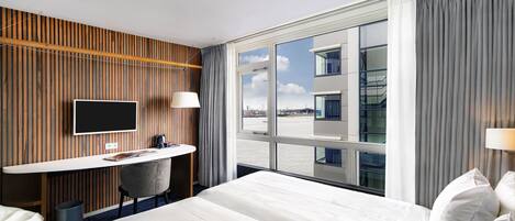 A hotel room with a large bed, a desk with a chair, a TV, and a view of the city through large windows.