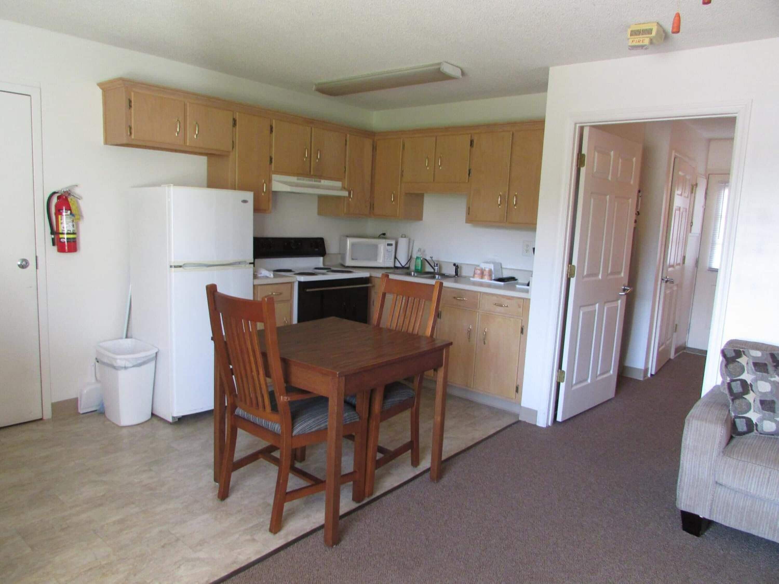 A compact kitchen with wooden cabinets, a white refrigerator, a stove, and a microwave. There's a dining table with chairs, a trash bin, and a fire extinguisher on the wall.