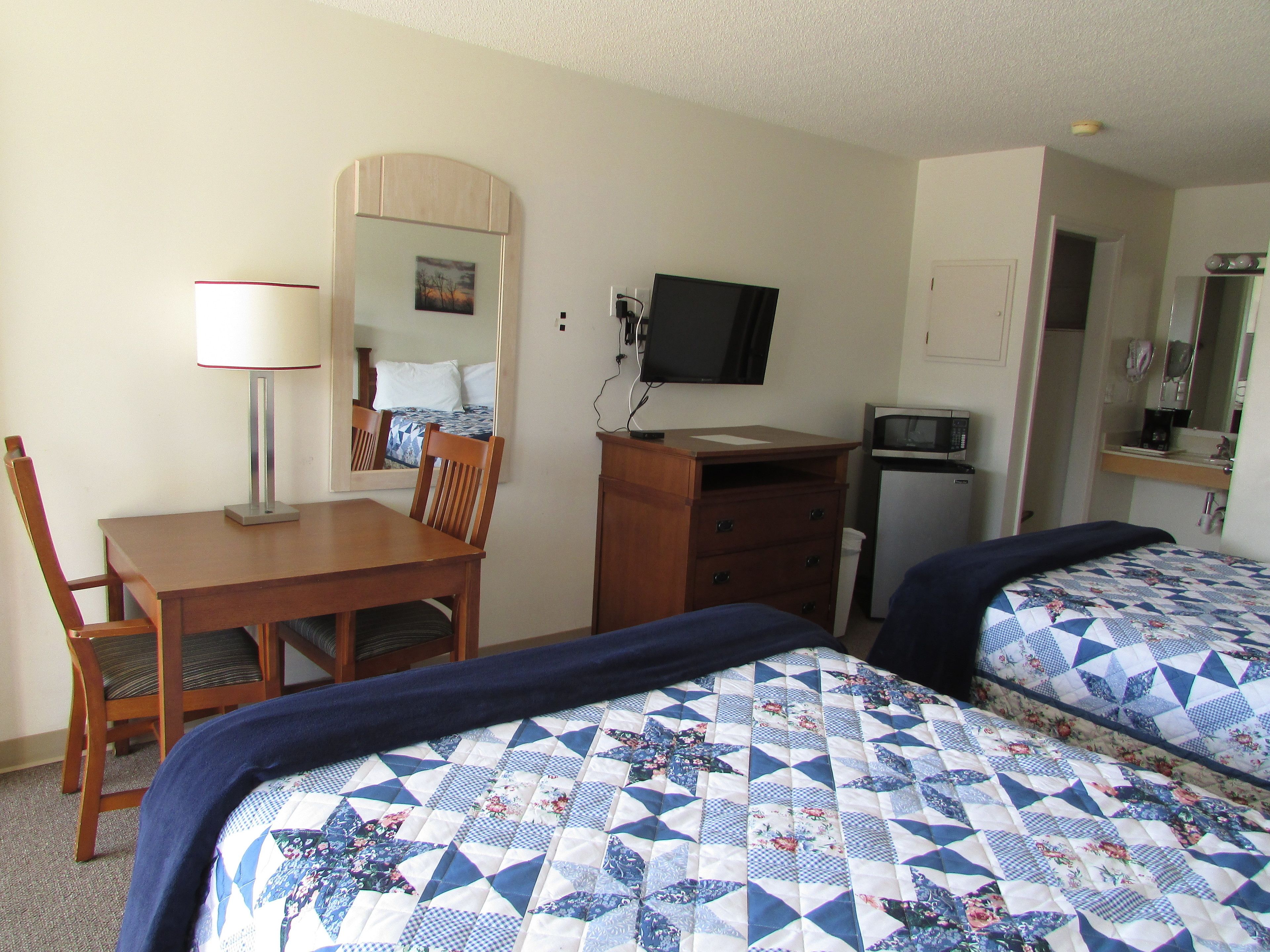 A hotel room with two beds, a desk with a chair, a lamp, a television, a microwave, and a dresser.