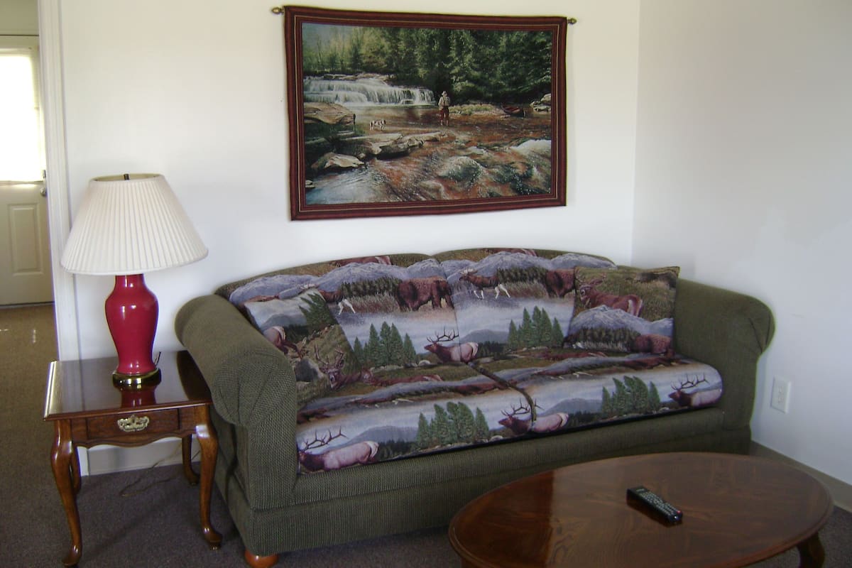 A living room with a green sofa, a red lamp, a painting of a forest scene, and a remote control on a coffee table.