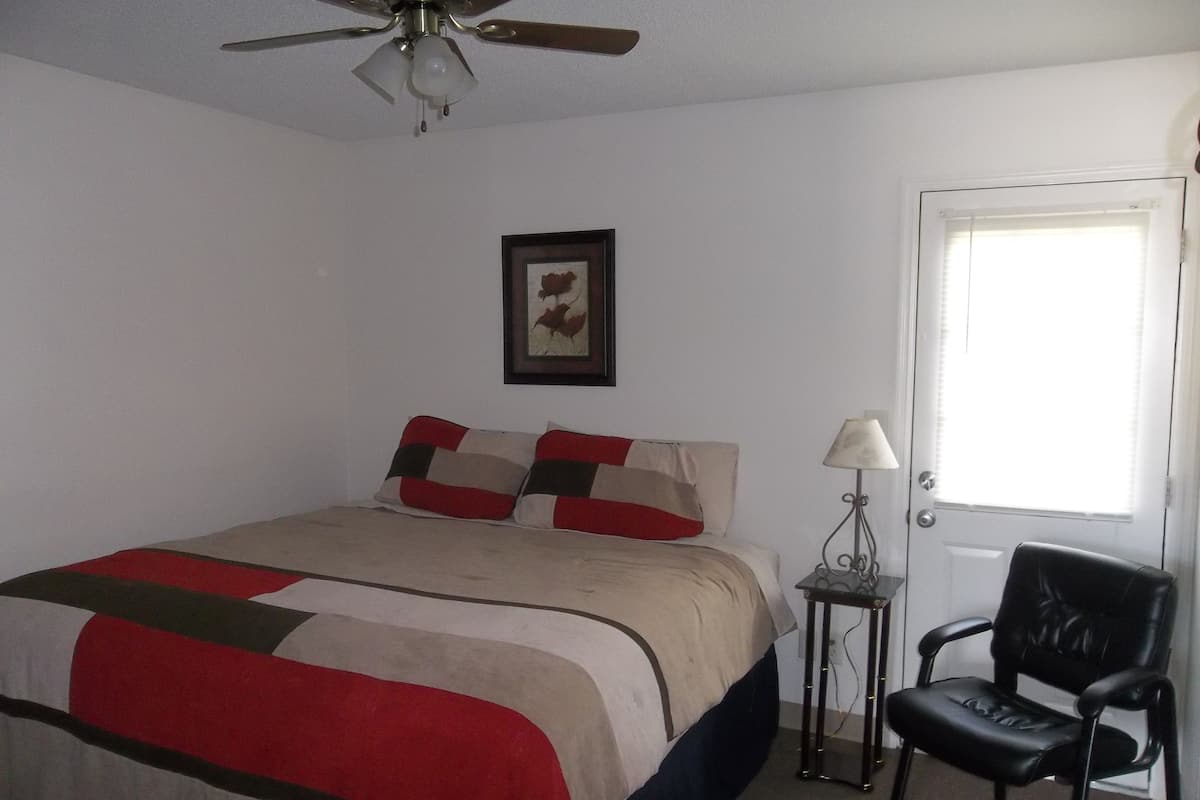 A bedroom with a bed, a ceiling fan, a framed picture on the wall, a door, and a chair.