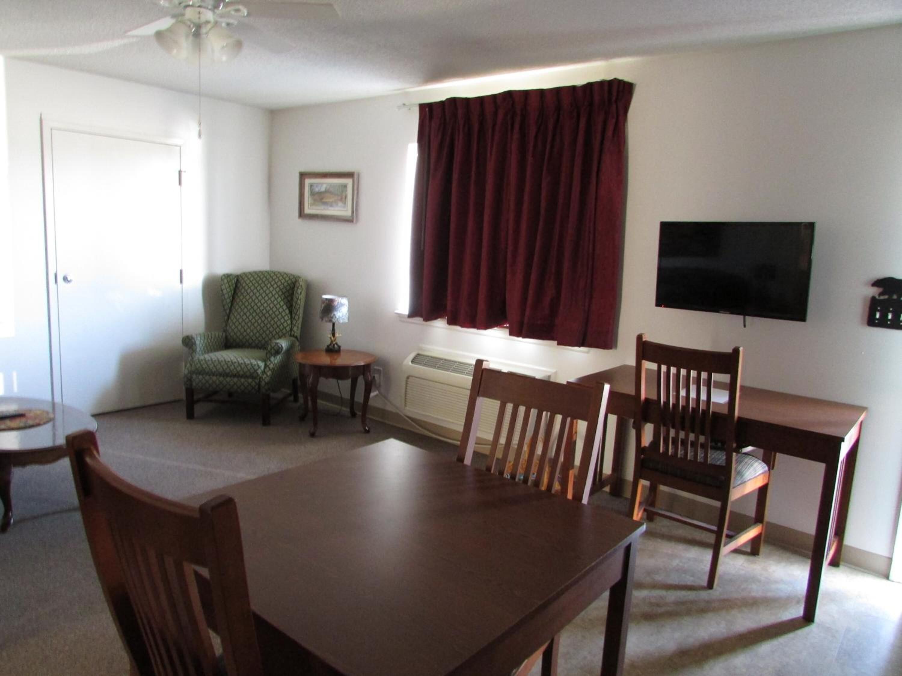 A living room with a dining table, two chairs, a green armchair, a small side table with a lamp, a window with curtains, a ceiling fan, and a TV mounted on the wall.