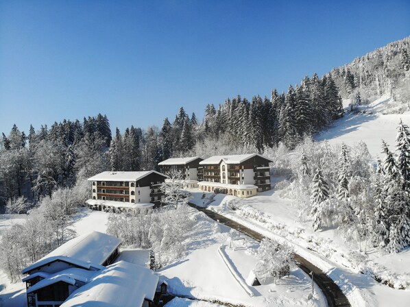 Un resort cubierto de nieve con varios edificios, incluyendo un gran albergue y cabañas más pequeñas, rodeado de un bosque denso y una pista de esquí.