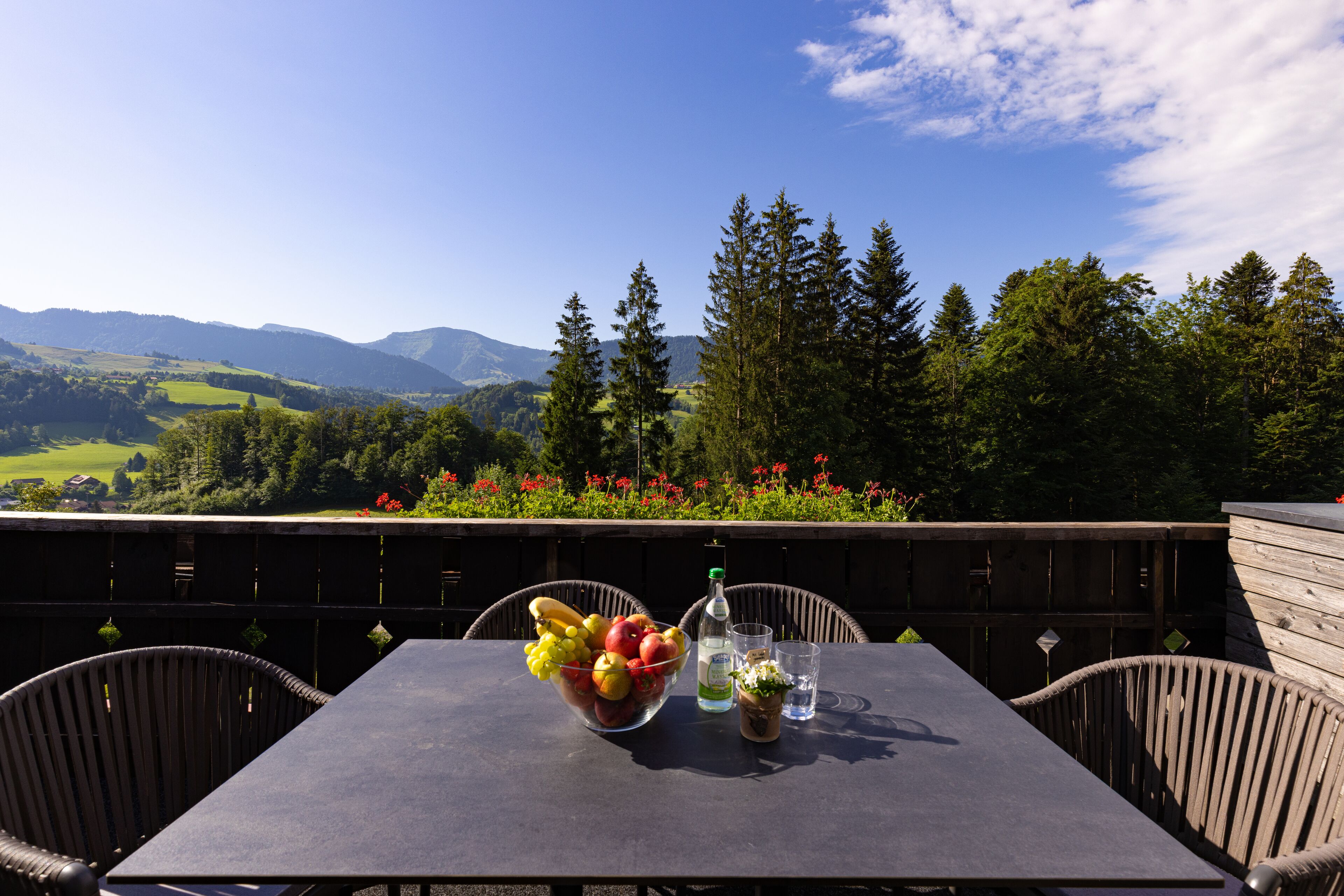 A table with a fruit bowl, a bottle, and glasses, set against a scenic mountain backdrop.