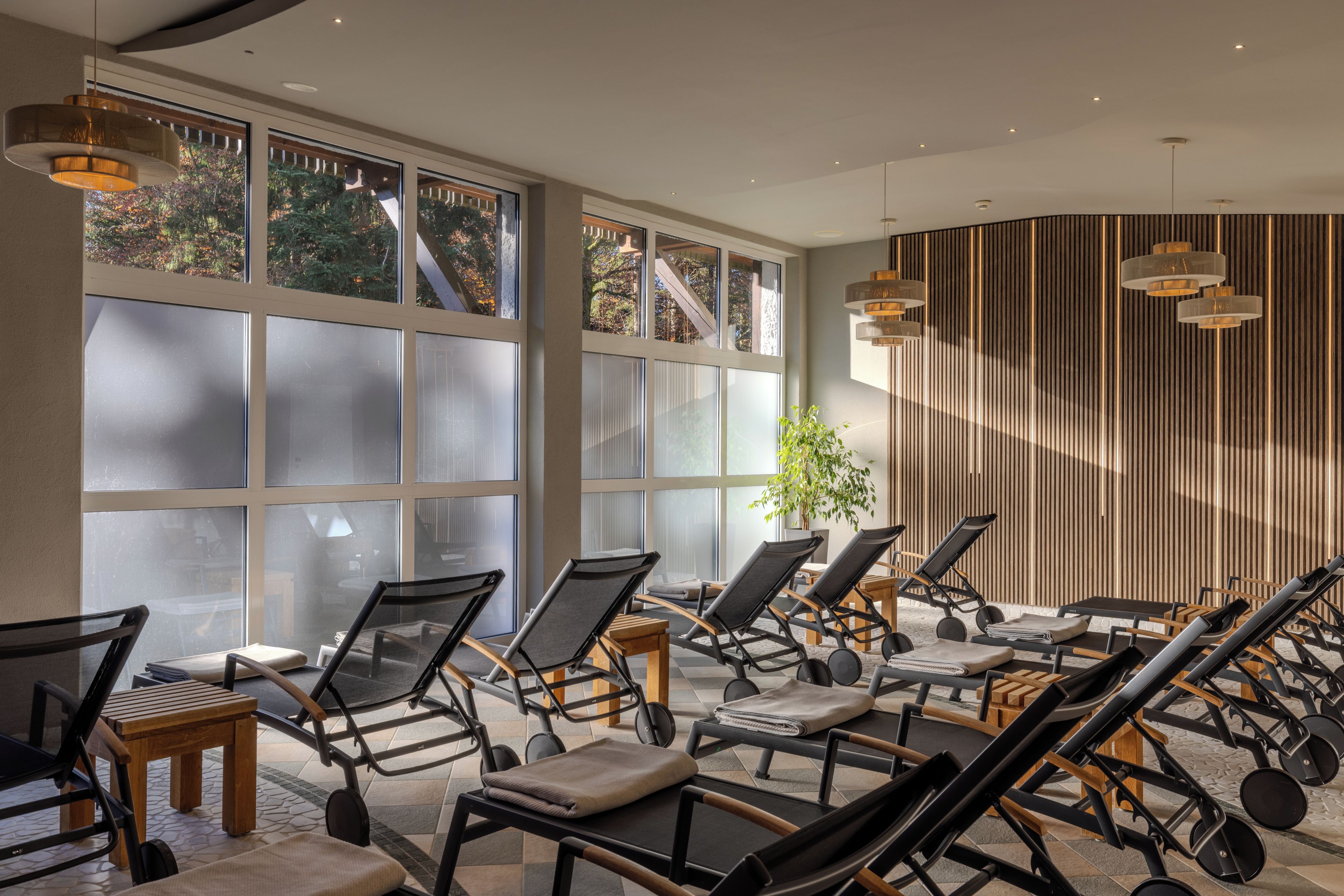 A sunlit room with multiple lounge chairs and tables, large windows, and a wooden slat wall.