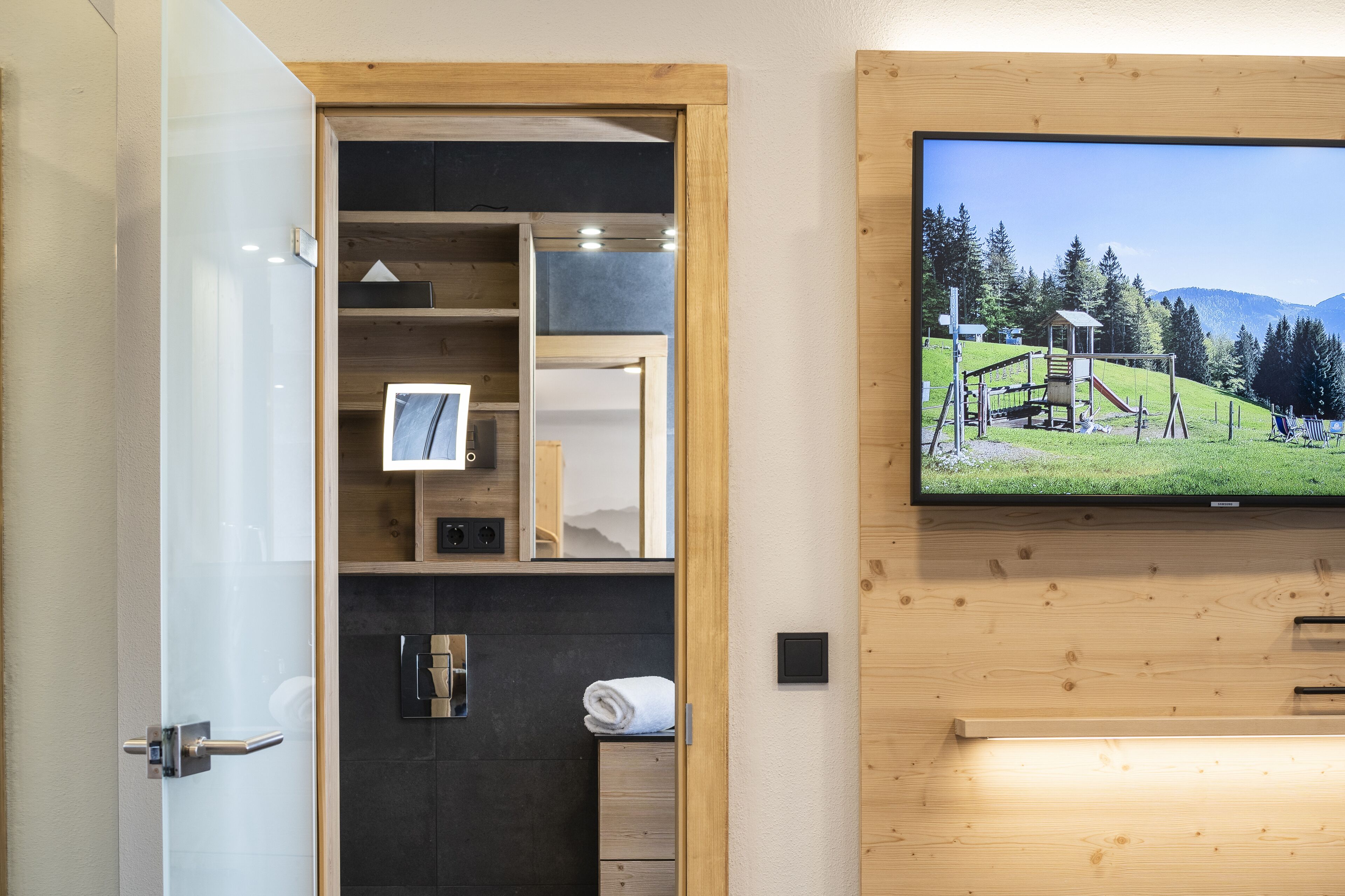 A modern bathroom with a wooden vanity, a built-in TV displaying a scenic landscape, and a towel rack.