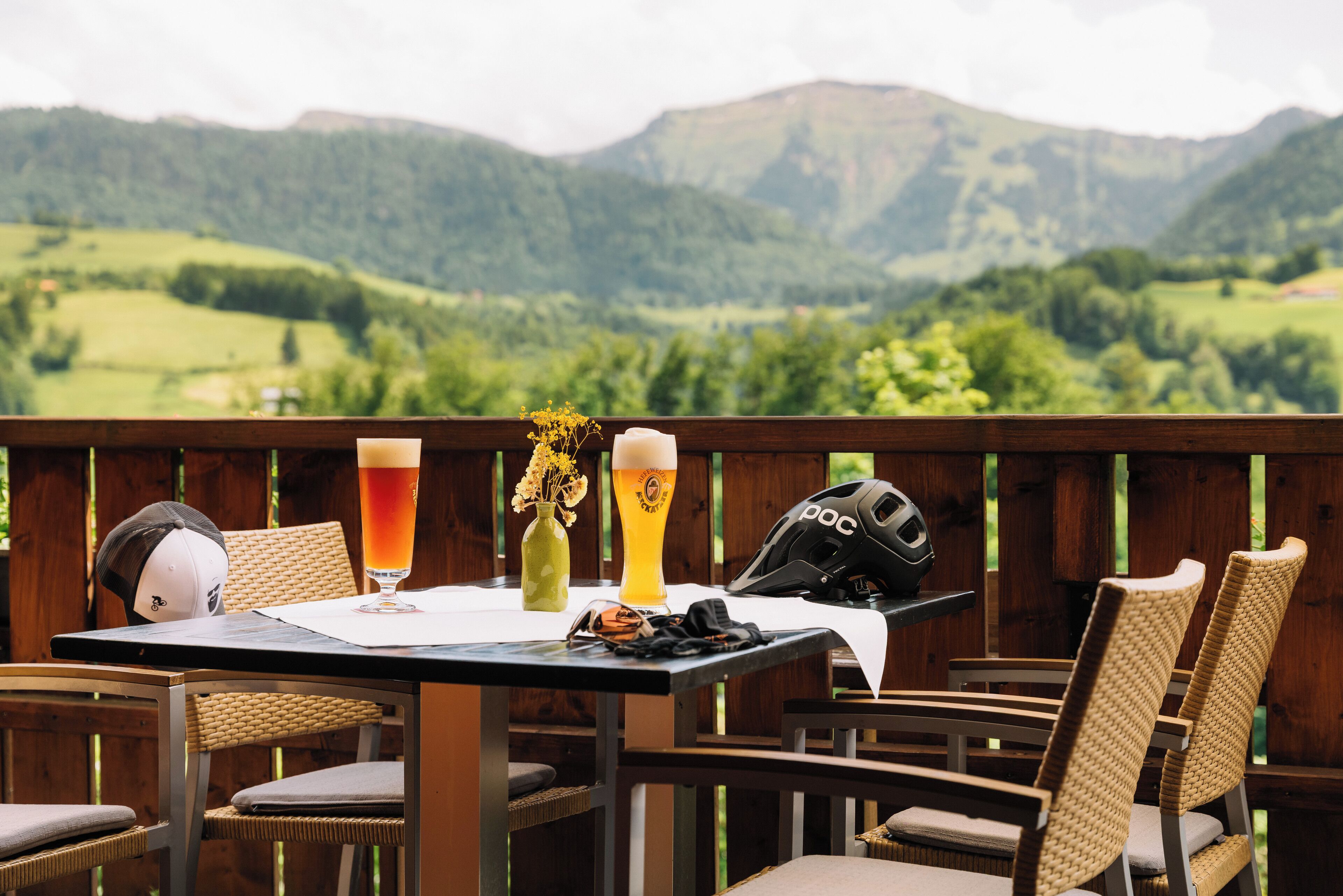 Een tafel met twee bierglazen, een helm en een helmzak op een houten terras, met uitzicht op een berg.