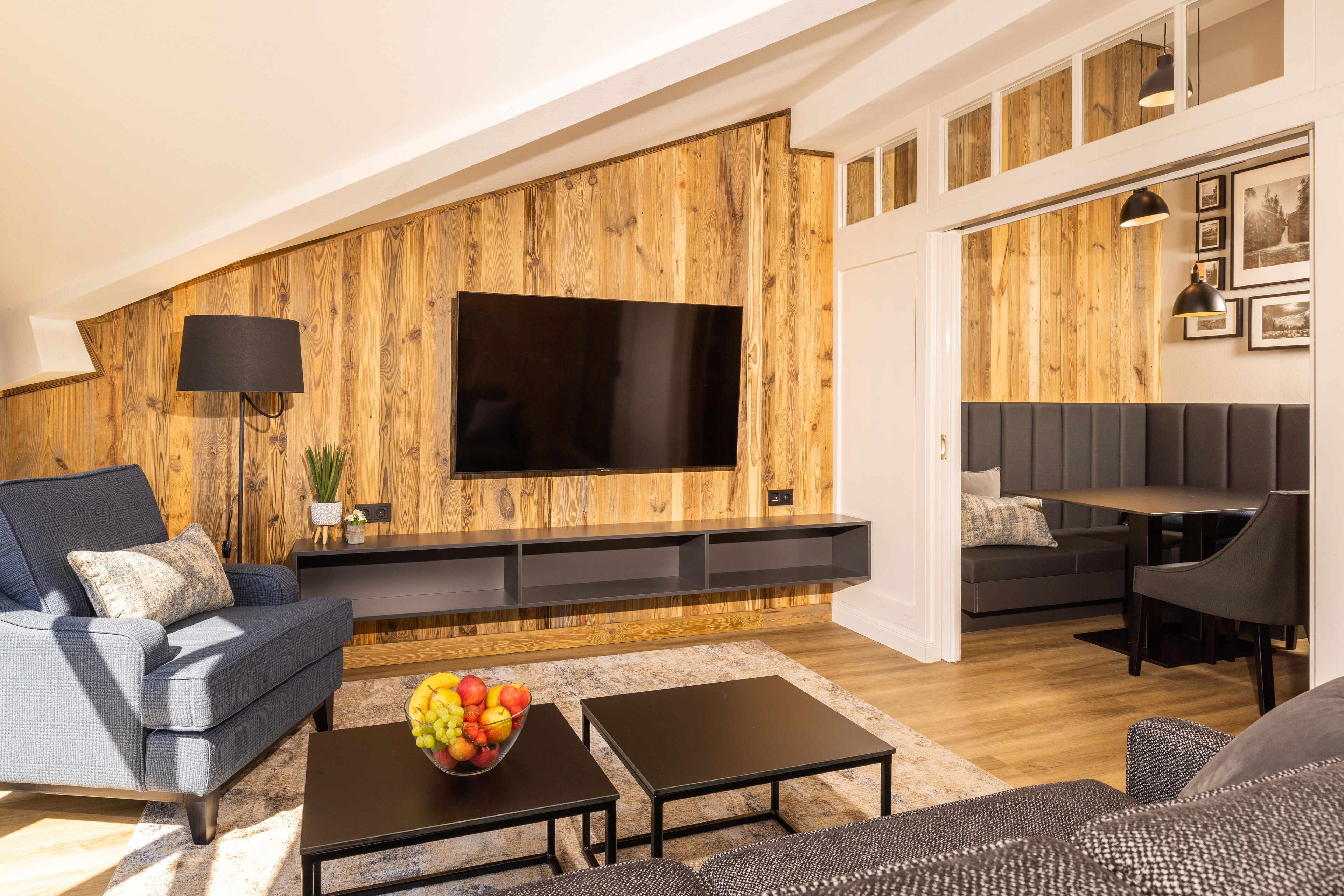Een moderne woonkamer met een houten accentmuur, een flatscreen-televisie, een bank, een salontafel met fruit en een eethoek.