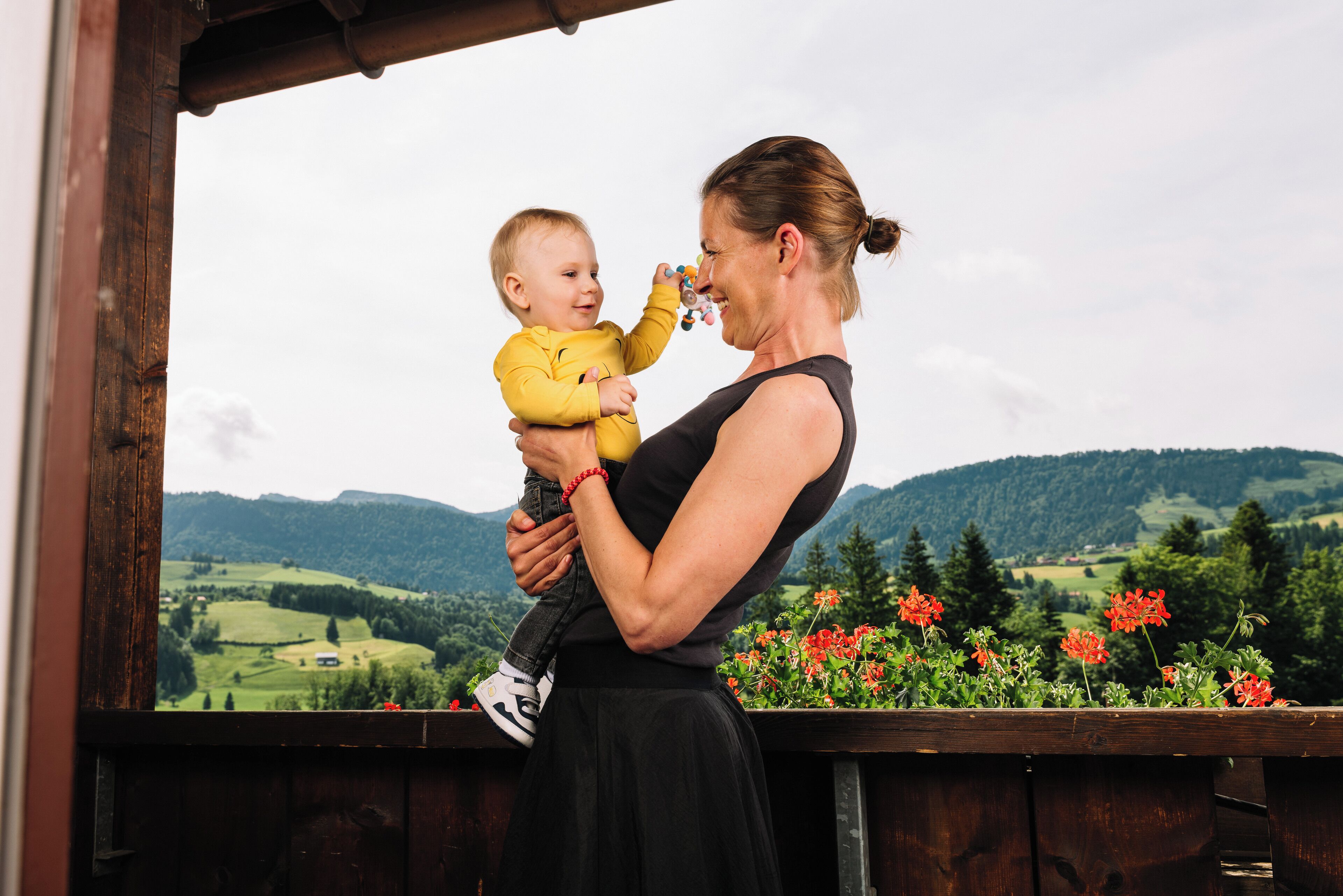 Una donna tiene in braccio un bambino su un balcone con vista su un paesaggio verde e delle colline.