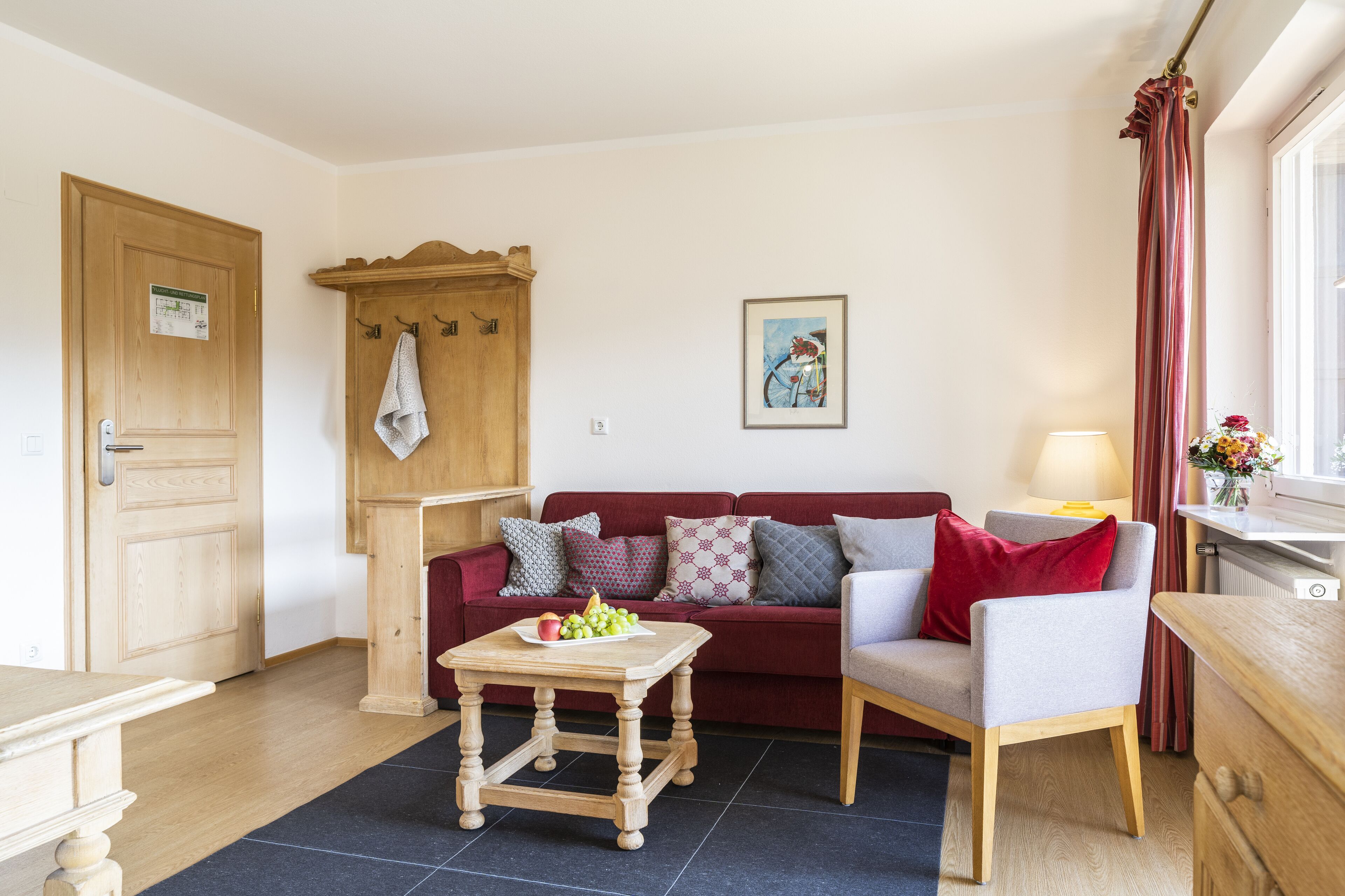 Una sala de estar acogedora con una puerta de madera, un sofá rojo con cojines estampados, una mesa de centro de madera con fruta y un sillón gris.