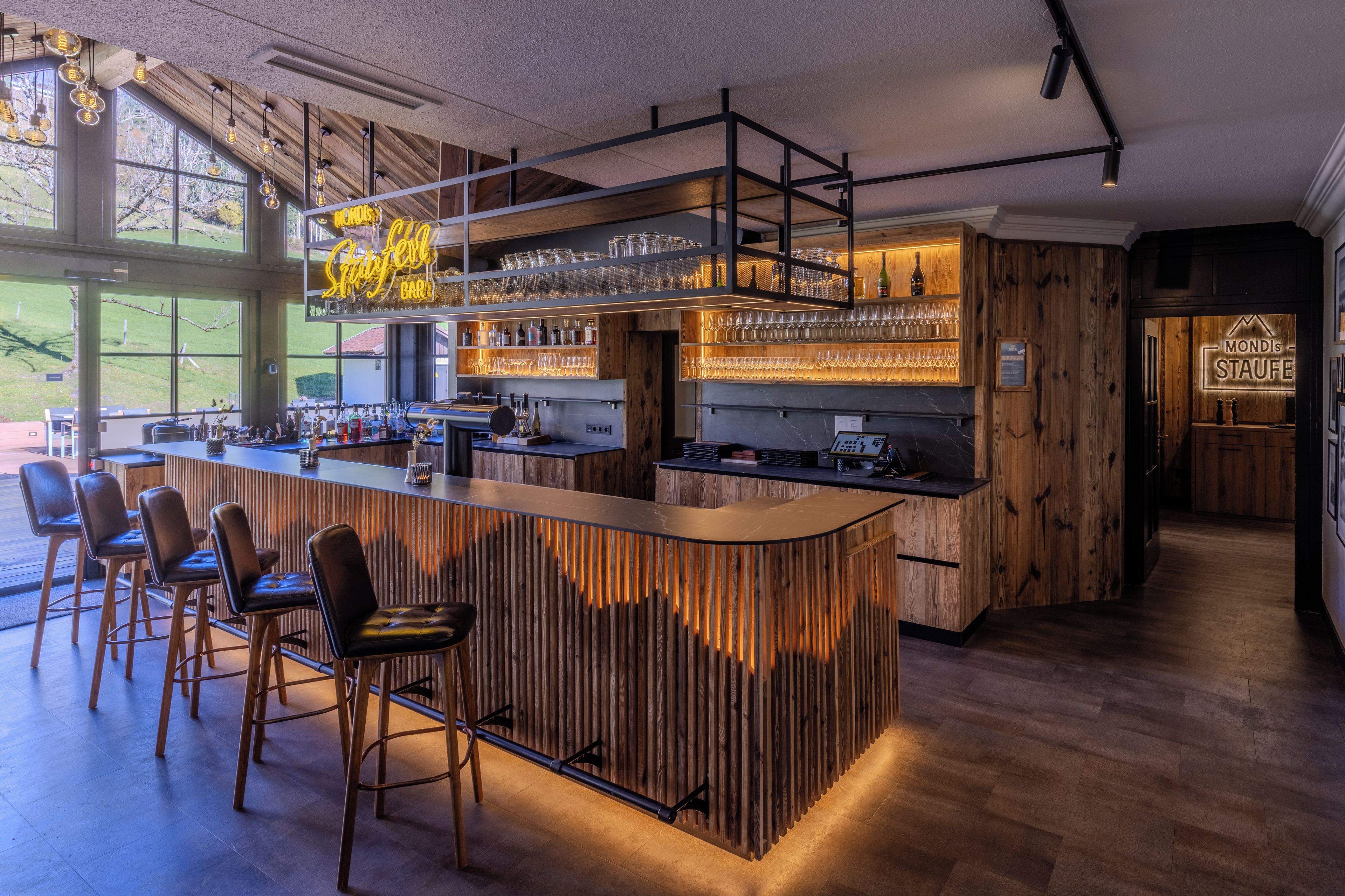Zona de bar con mostrador de madera, taburetes y vistas al exterior a través de amplios ventanales.