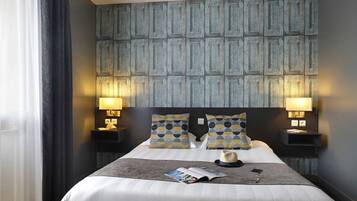 A hotel room with a large bed, two bedside tables with lamps, patterned pillows, a magazine, and a hat on the bed.