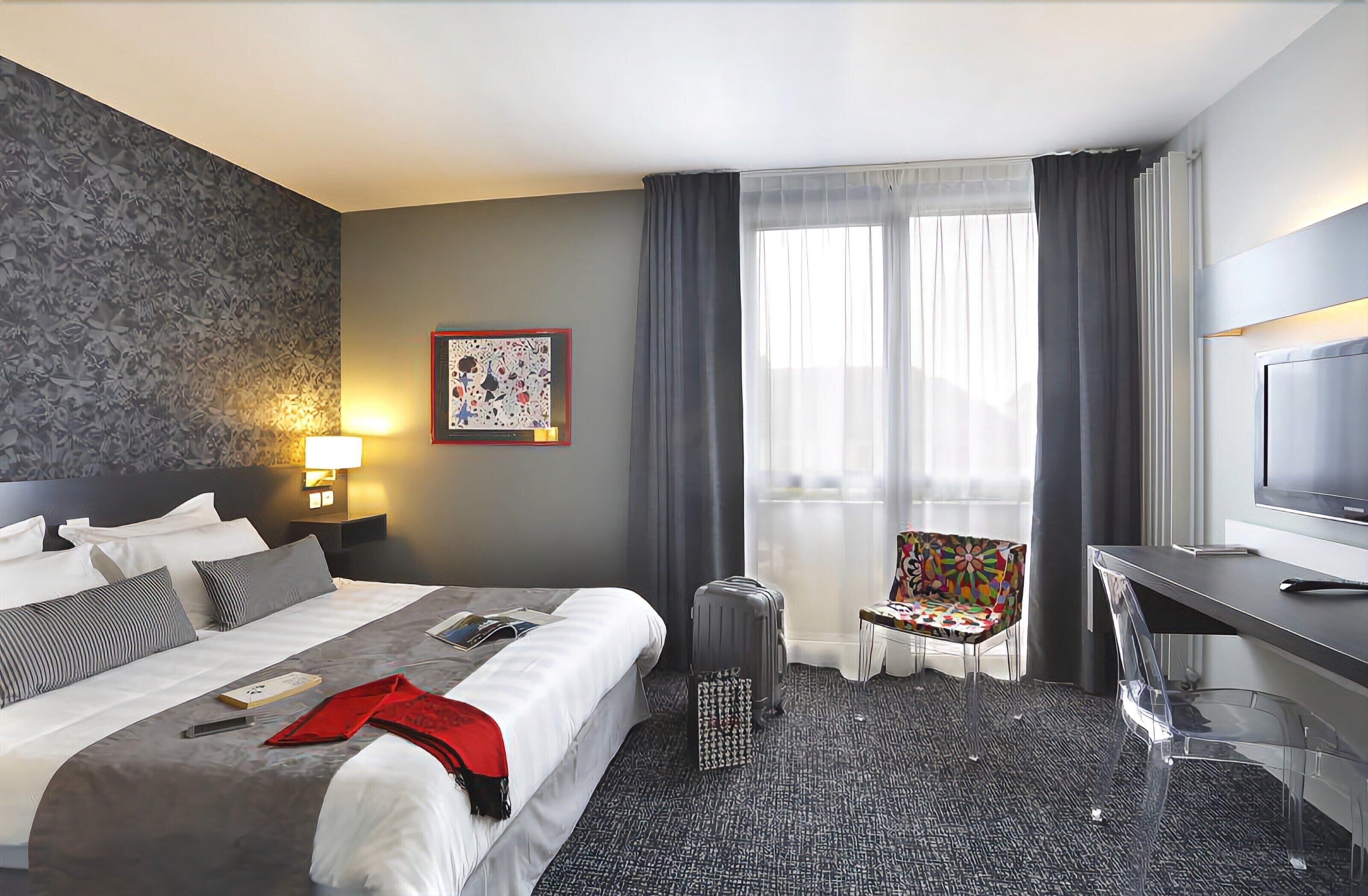 Una habitación de hotel con cama, escritorio, silla, lámpara y ventana con cortinas.