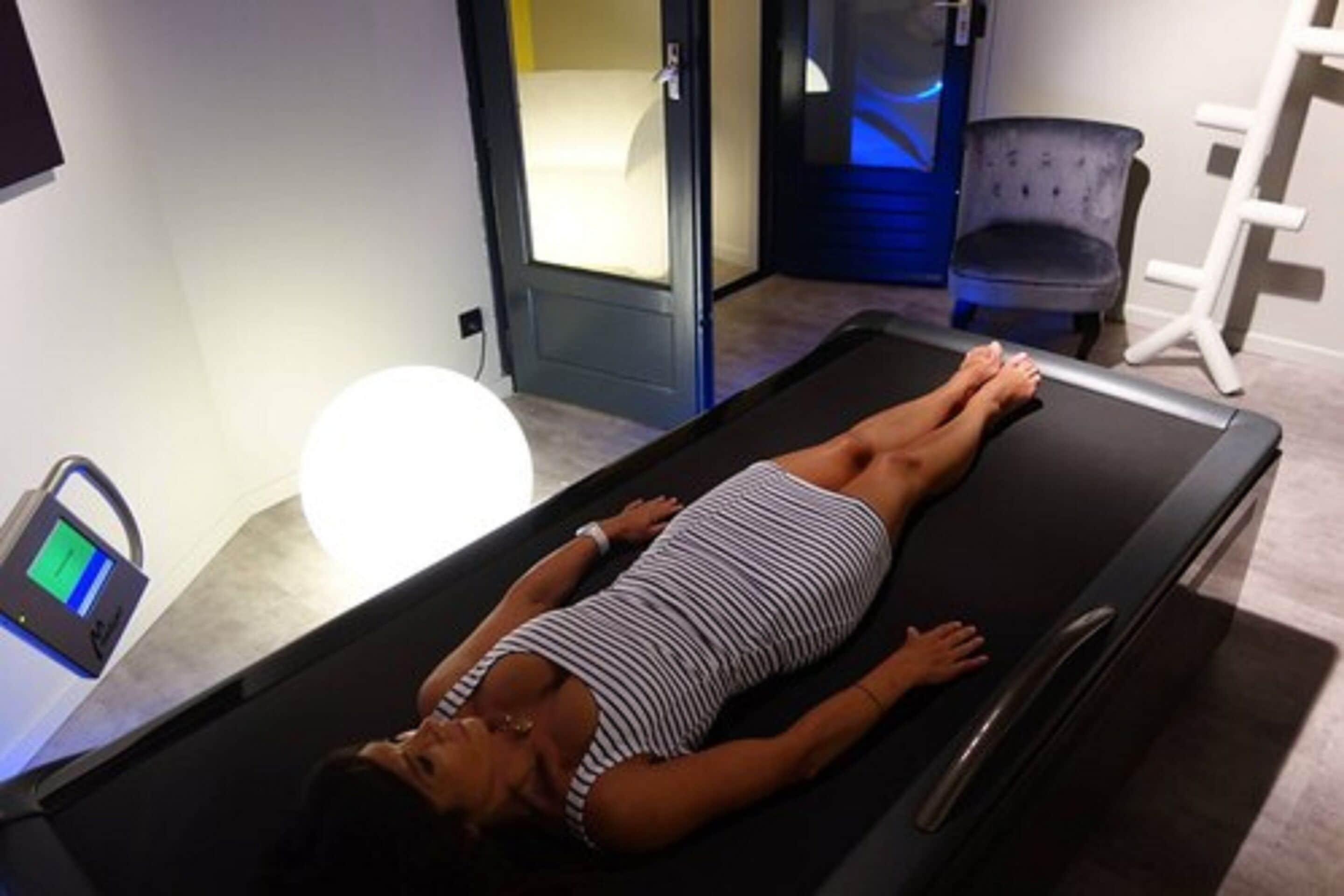 A woman lying on a massage table in a room with a massage chair and a digital device on a table.