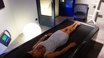 A woman lying on a massage table in a room with a massage chair and a digital device on a table.