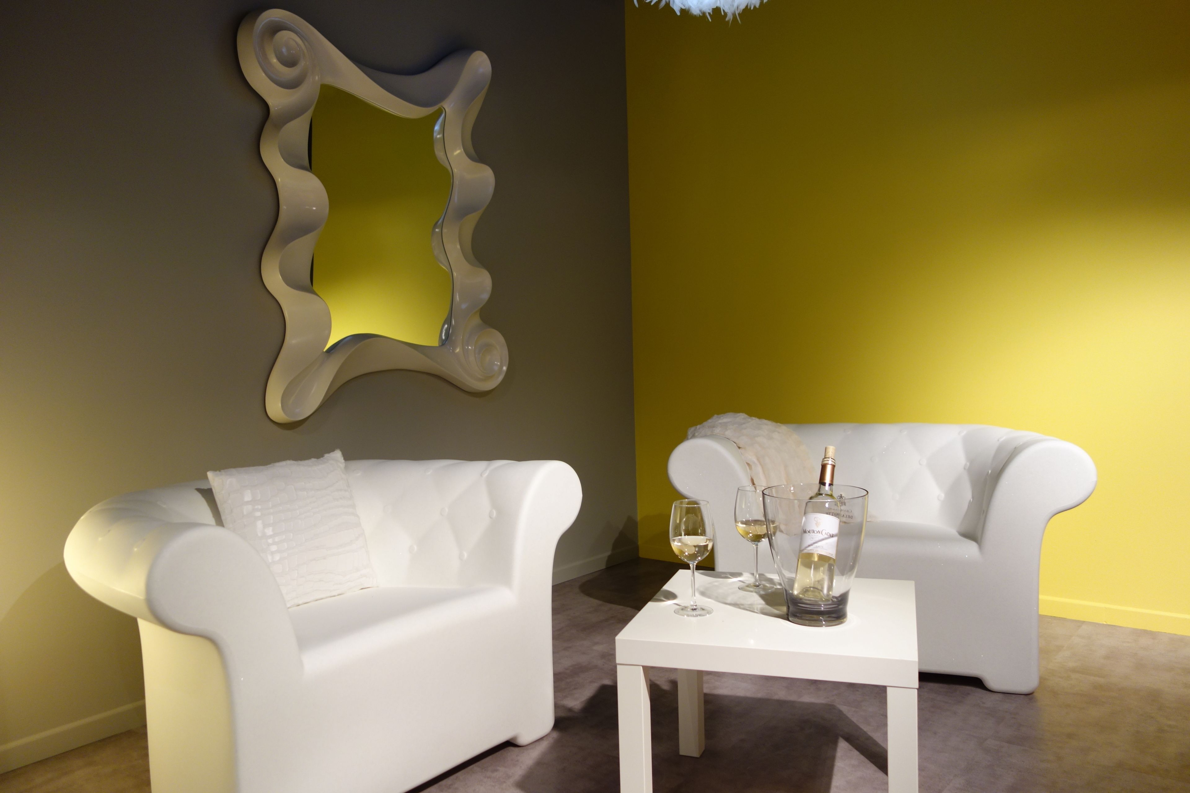Una habitación moderna con dos sofás blancos, una mesa blanca con vasos y una botella, y un espejo decorativo en la pared.