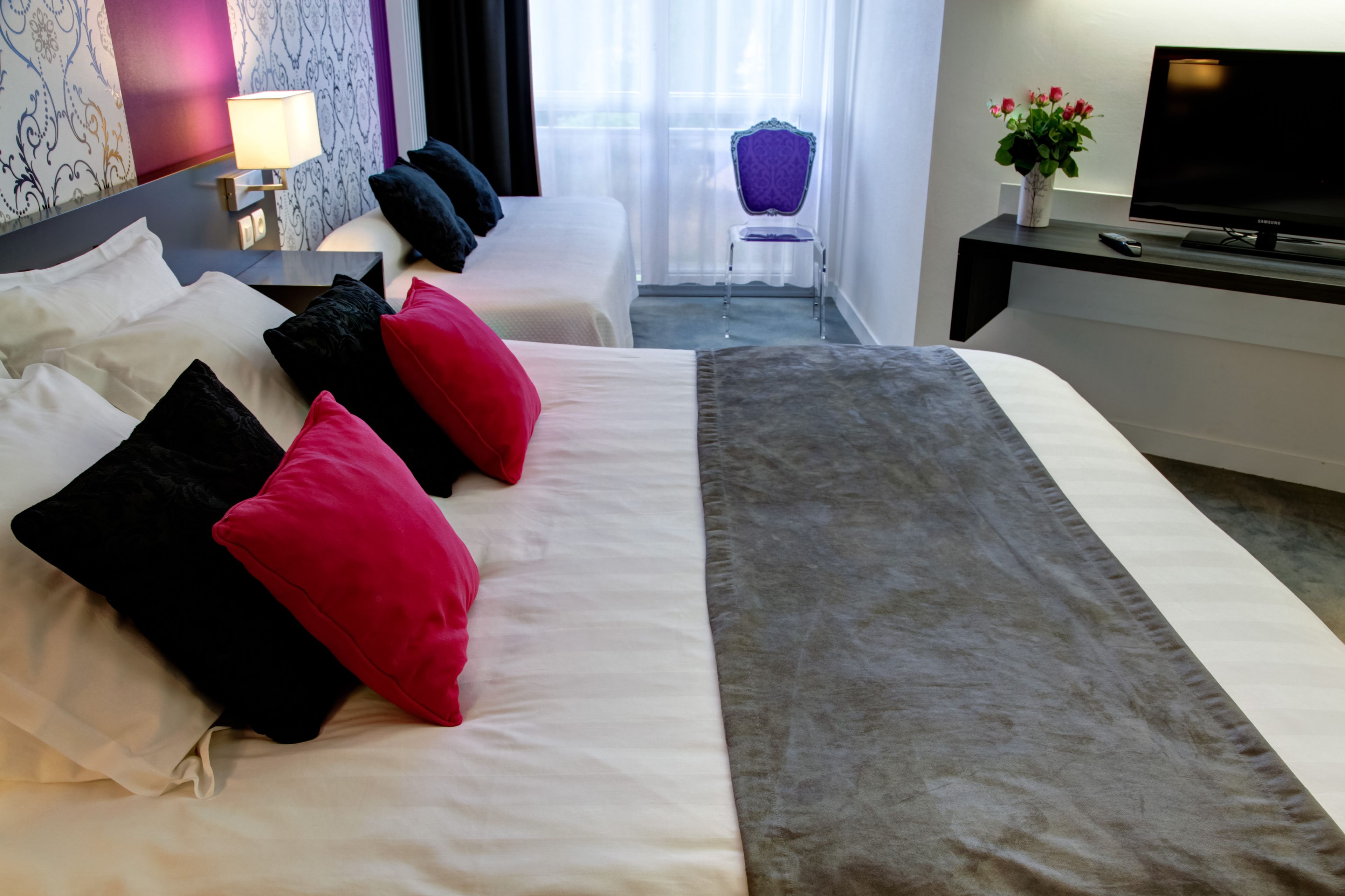 A hotel room with a large bed, multiple pillows, a TV, a chair, and a small table with a lamp.
