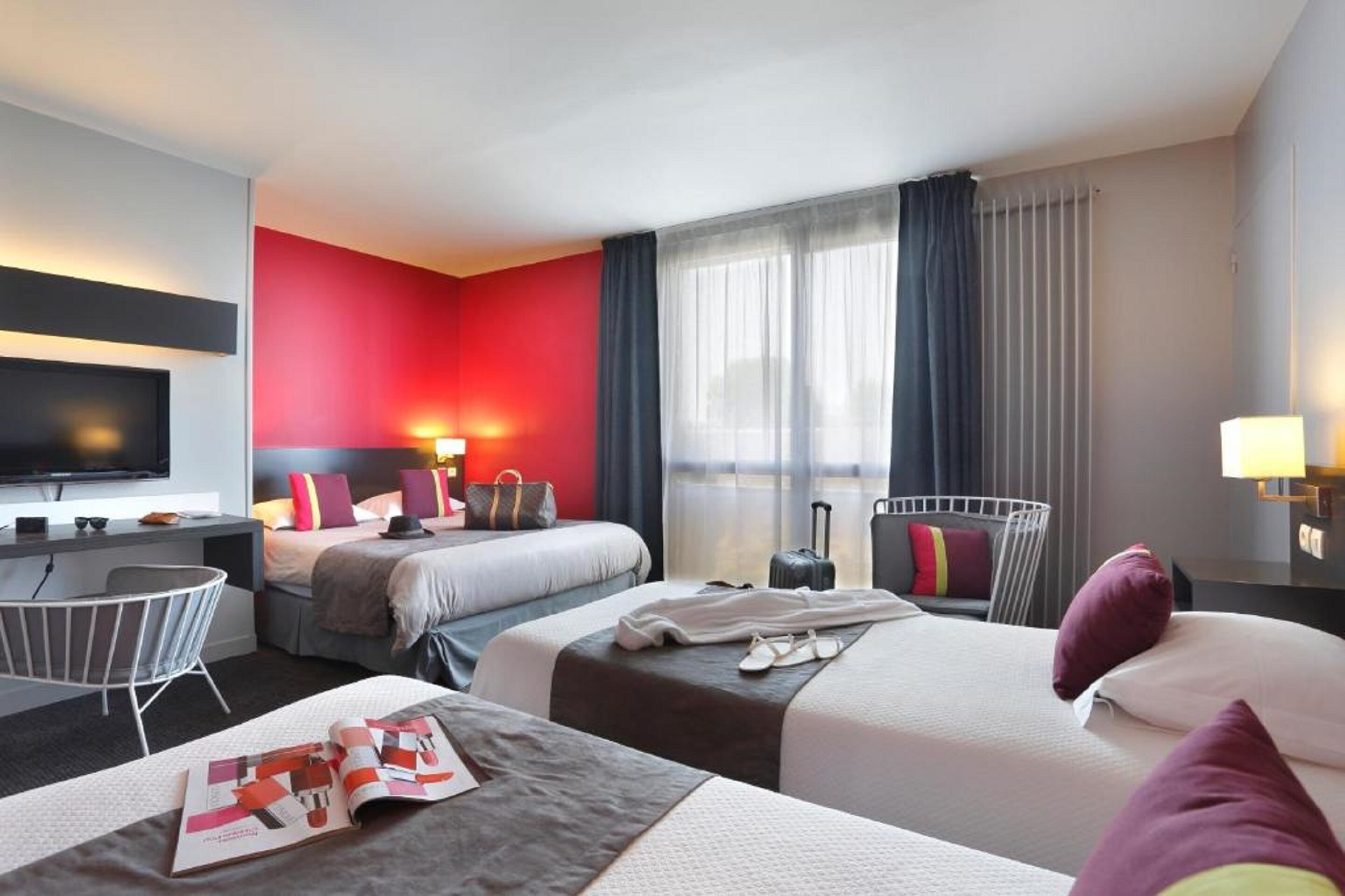 A hotel room with two beds, a desk, a TV, and a window with curtains.