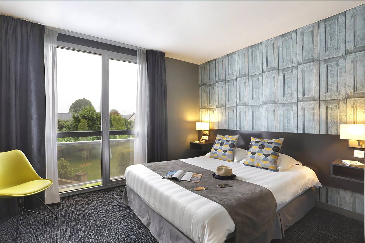 Une chambre d’hôtel avec un grand lit, une chaise jaune, une vue à travers une porte coulissante et un mur à texture.