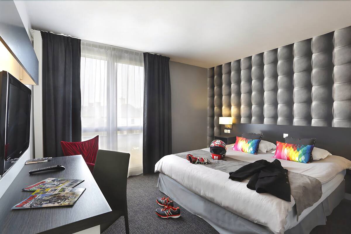 Une chambre d’hôtel avec un lit, un bureau avec une télévision, une chaise et une fenêtre avec des rideaux.
