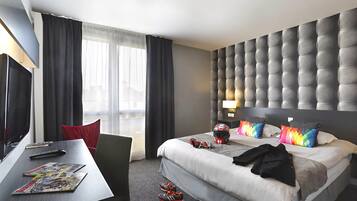 A hotel room with a bed, a desk with a TV, a chair, and a window with curtains.
