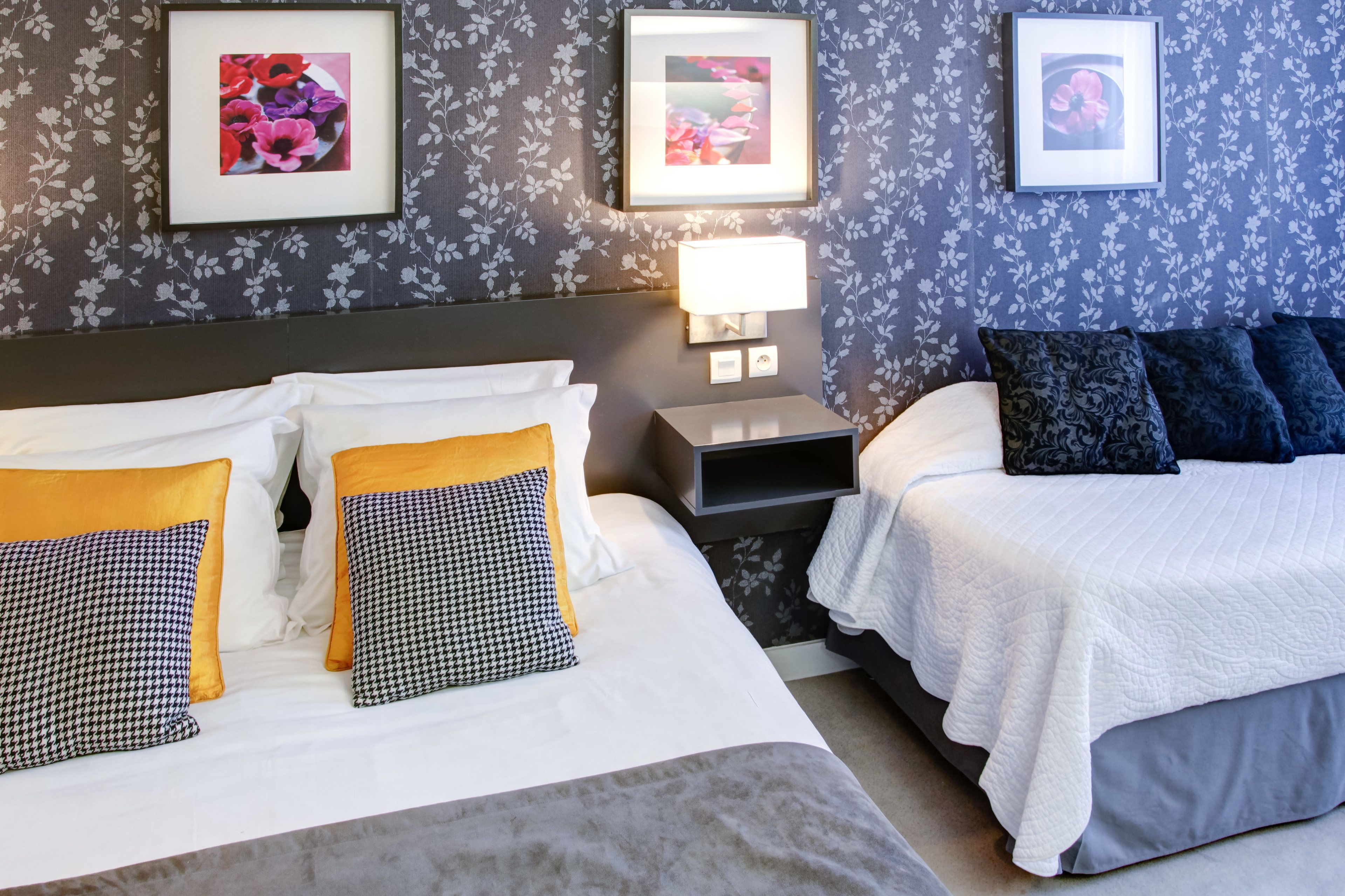 A hotel room with two beds, a nightstand with a lamp, and floral wallpaper.